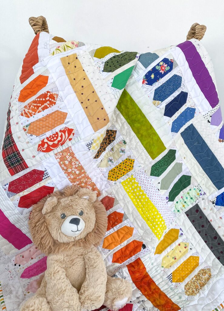 Ranbow baby Prismatic Abacus Quilt is draped over a wicker basket in front of a white wall and a baby lion toy sits on top of the quilt