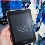 Amazon Kindle Paperwhite in front of a colorful quilt. The Kindle is showing a custom black and white lock screen that says, "I read because I can't take my sewing machine".